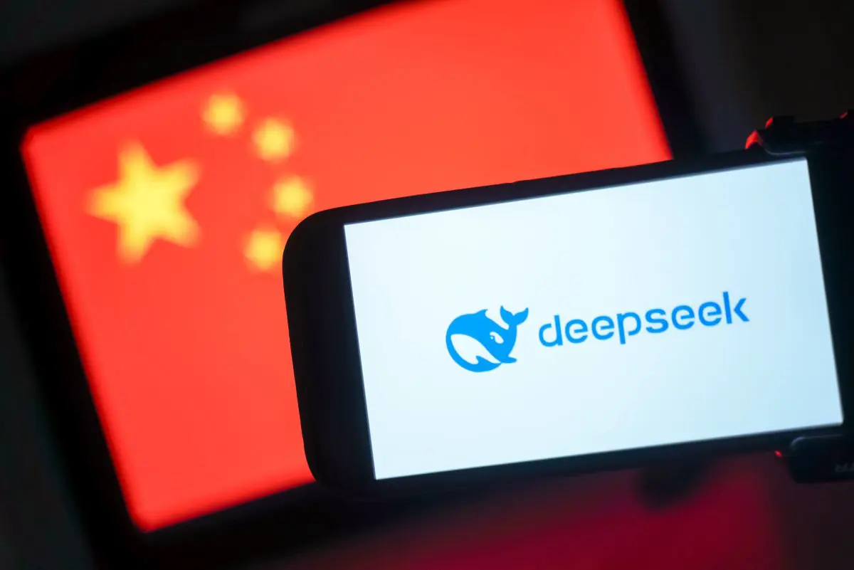 تظهر الصورة هاتفاً عليه شعار تطبيق DeepSeekعلى الشاشة وفي الخلفية علم الصين.